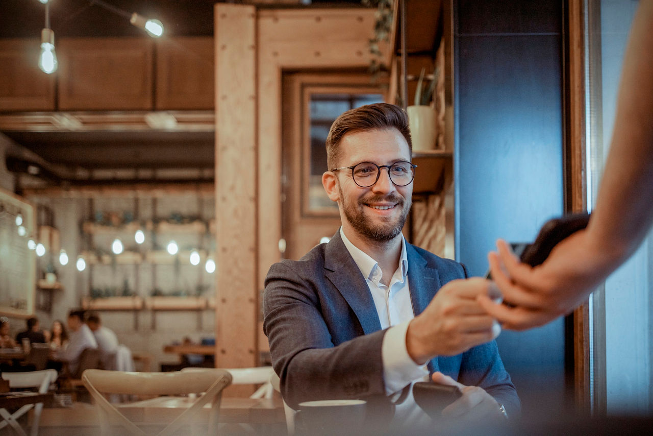 Ein junger Mann mit Brille sitzt im lockeren Business Outfit in einem Restaurant und bezahlt gerade mit Karte. Elegant Male DresseEin junger Mann mit Brille sitzt im lockeren Business Outfit in einem Restaurant und bezahlt gerade mit Karte.d in a Suit Using Credit Card on Pos Terminal in Cafeteria