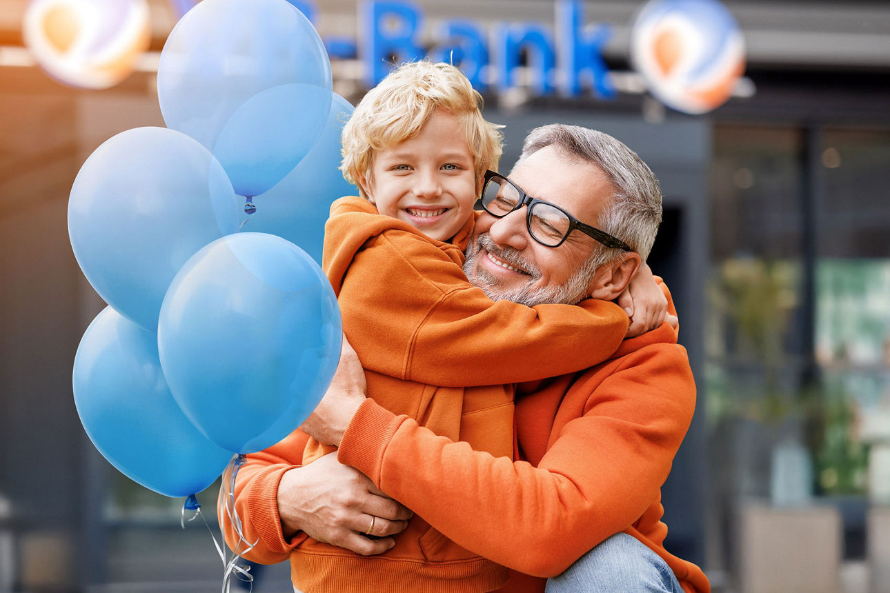 Vater mit Kind auf dem Arm und blauen Luftballons vor dem Bankgebäude