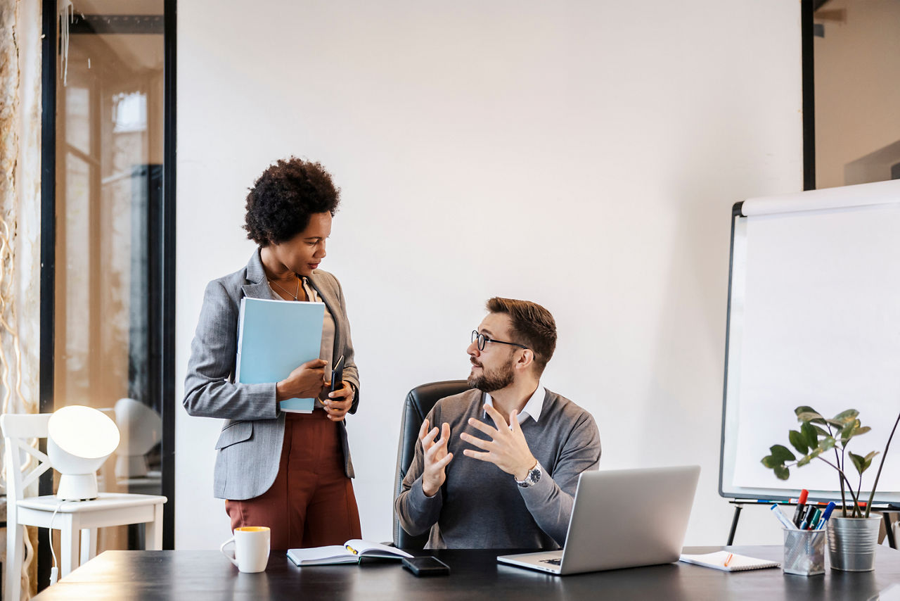Zwei Geschäftspersonen im Gespräch in einem modernen Büro mit Laptop, Unterlagen und Flipchart.