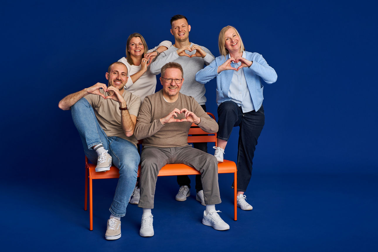 Eine Gruppe Mitarbeitenden bei einem Fotoshooting, auf orangener Bank, formen ein Herz mit den Händen. Auf blauem Hintergrund