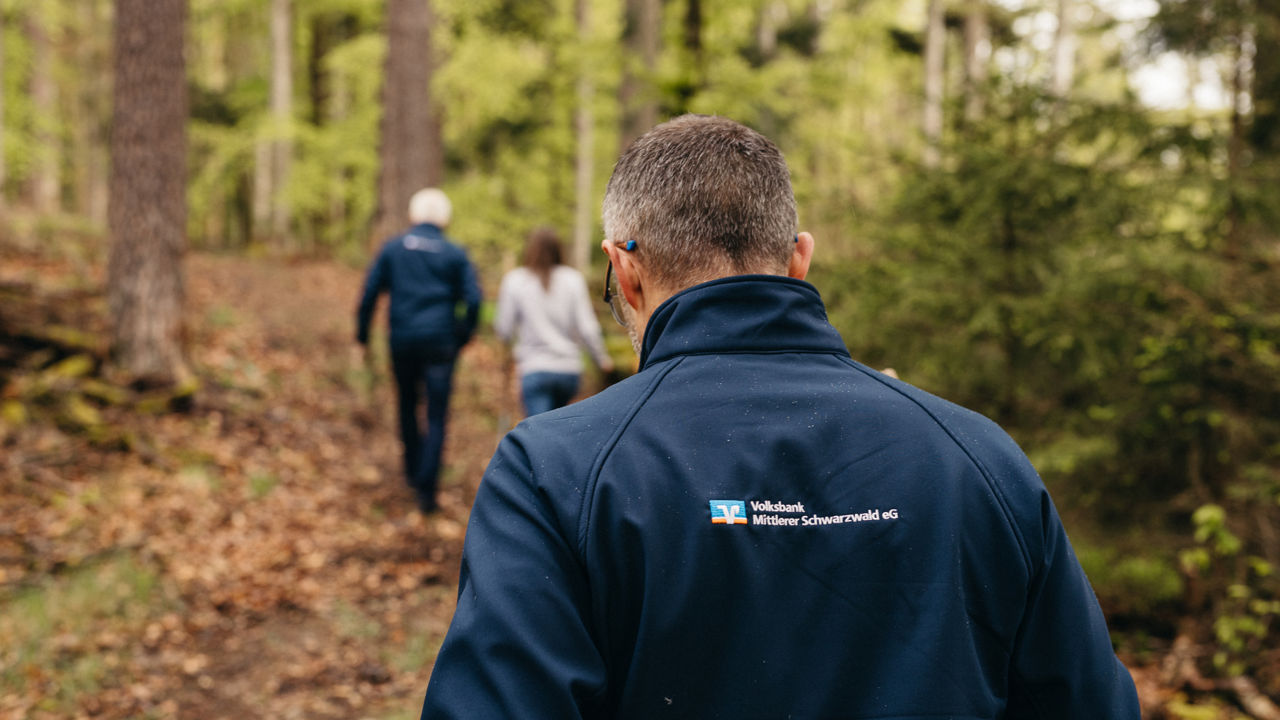 Vorstand spaziert durch den Wald. Dunkelblaue Softshell-Jacke mit Volksbank Logo, von hinten fotografiert.