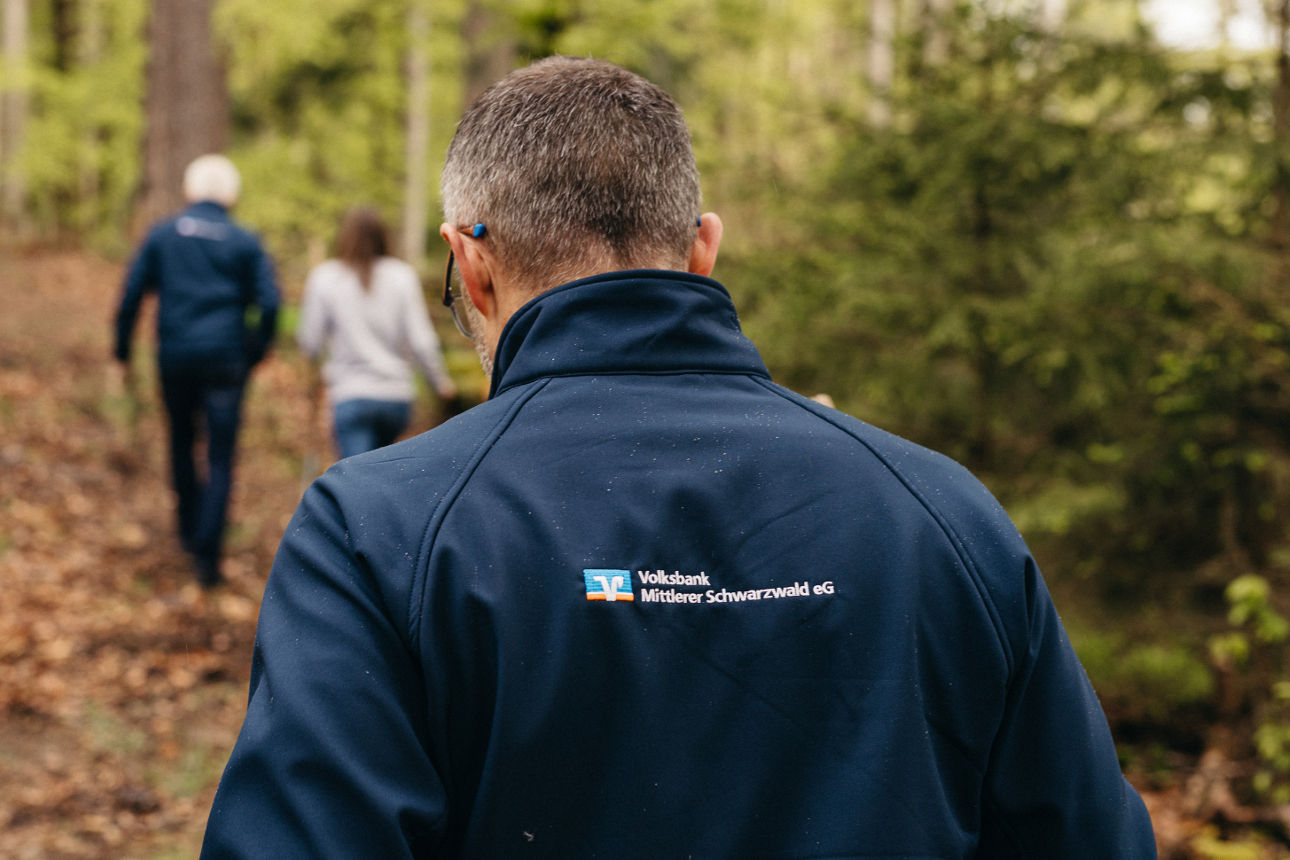 Vorstand spaziert durch den Wald. Dunkelblaue Softshell-Jacke mit Volksbank Logo, von hinten fotografiert.