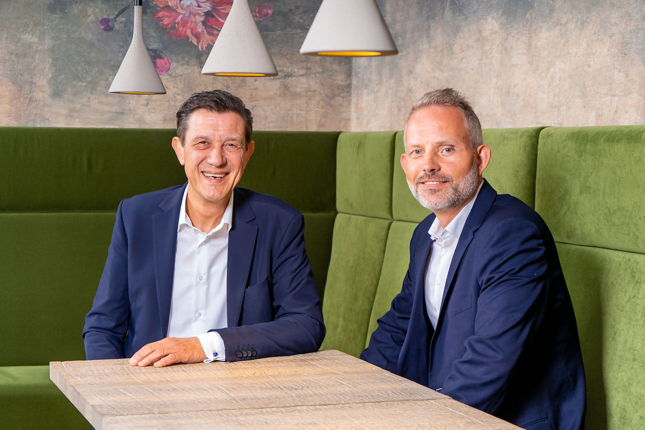 Zwei Männer in blauen Anzügen, die an einem Tisch auf einem grünen Sessel sitzen.