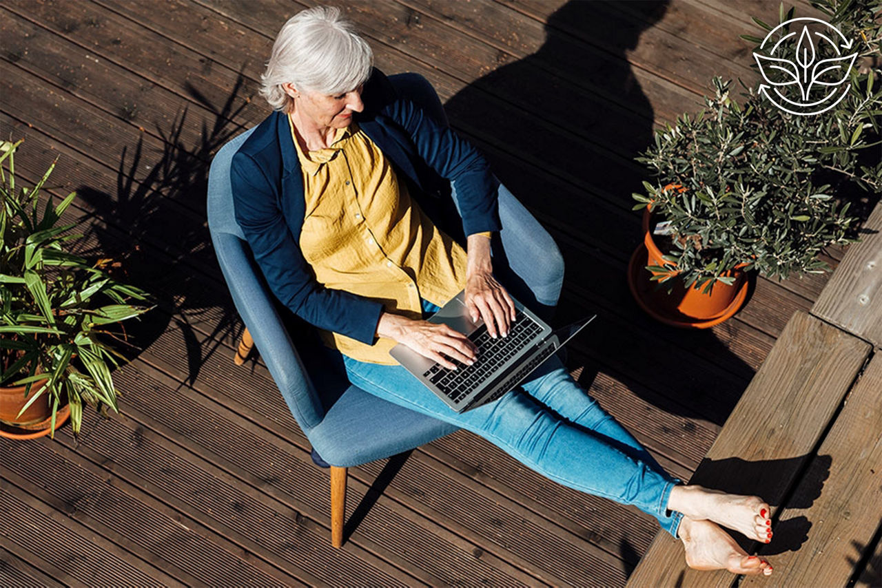 Eine ältere Frau sitzt entspannt auf einer Terrasse und arbeitet am Laptop