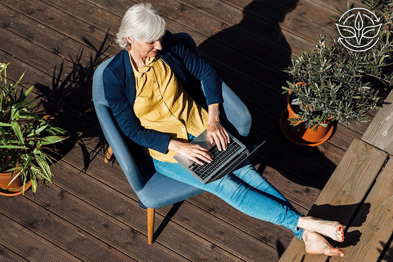 Eine ältere Frau sitzt entspannt auf einer Terrasse und arbeitet am Laptop