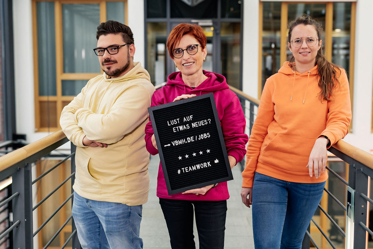 Kollegen stehen in der Bank und halten ein Schild hoch mit der Aufschrift ,,Lust auf etwas Neues?''