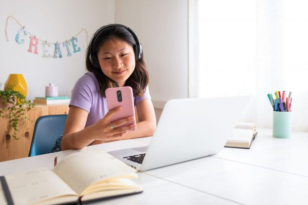 Mädchen lernt am Schreibtisch mit Laptop und Handy in der Hand