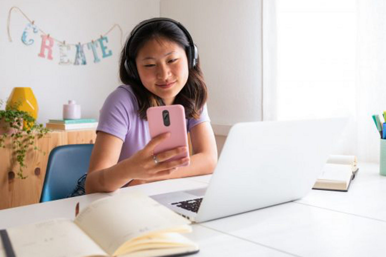Mädchen lernt am Schreibtisch mit Laptop und Handy in der Hand
