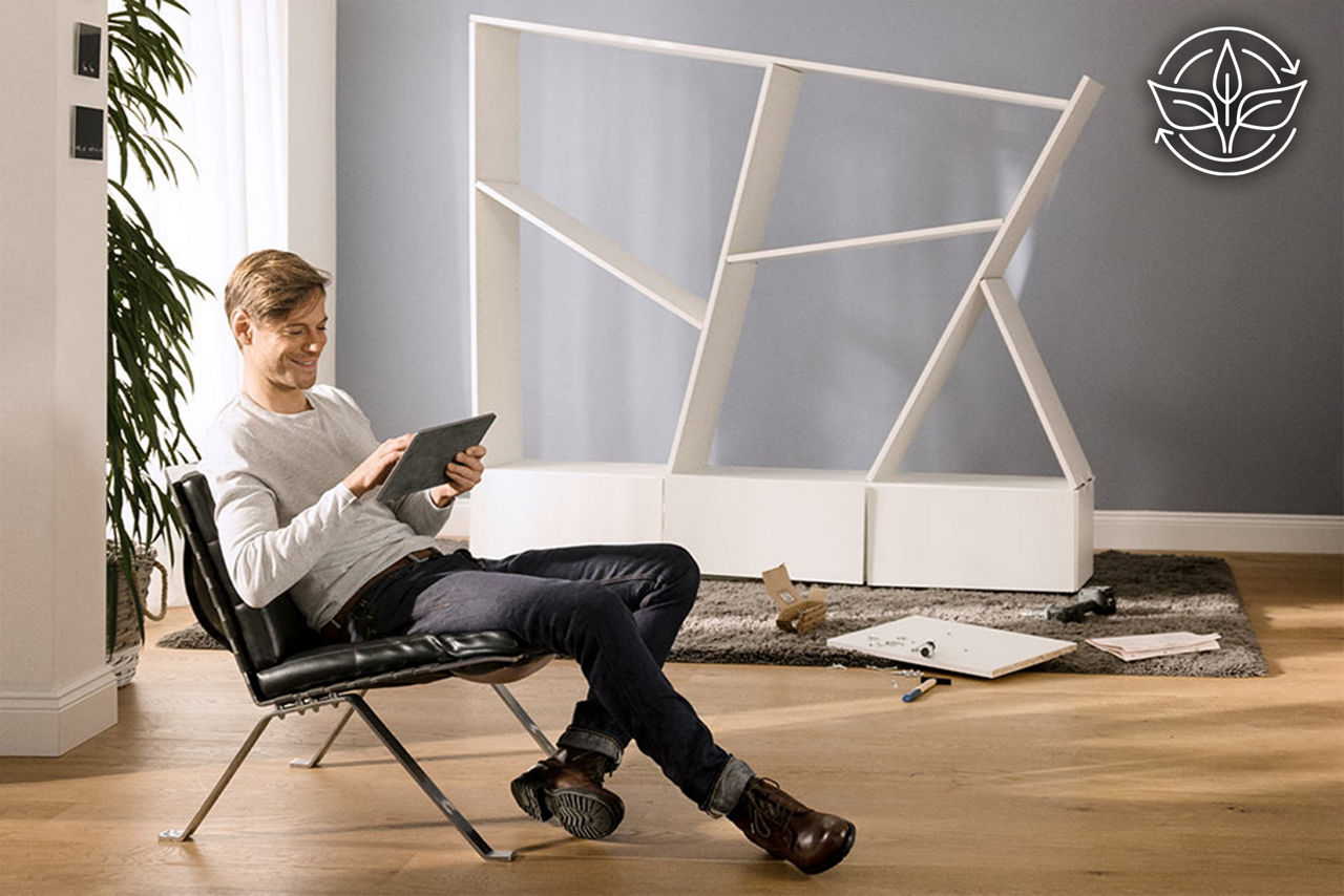 Ein Mann sitzt mit einem Tablet lächelnd vor einem unvollständig aufgebauten Bücherregal