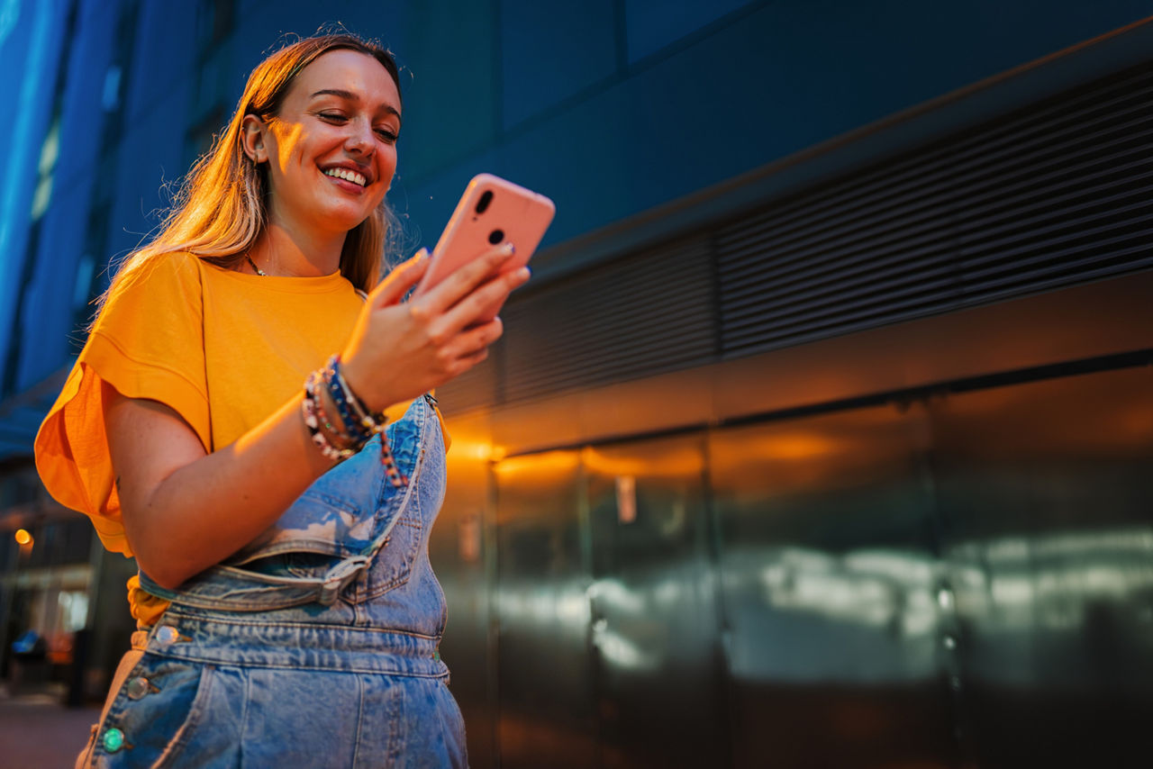Ein glückliches junges Mädchen benutzt ihr Smartphone, um im Internet zu surfen, während sie nachts auf der Straße spazieren 