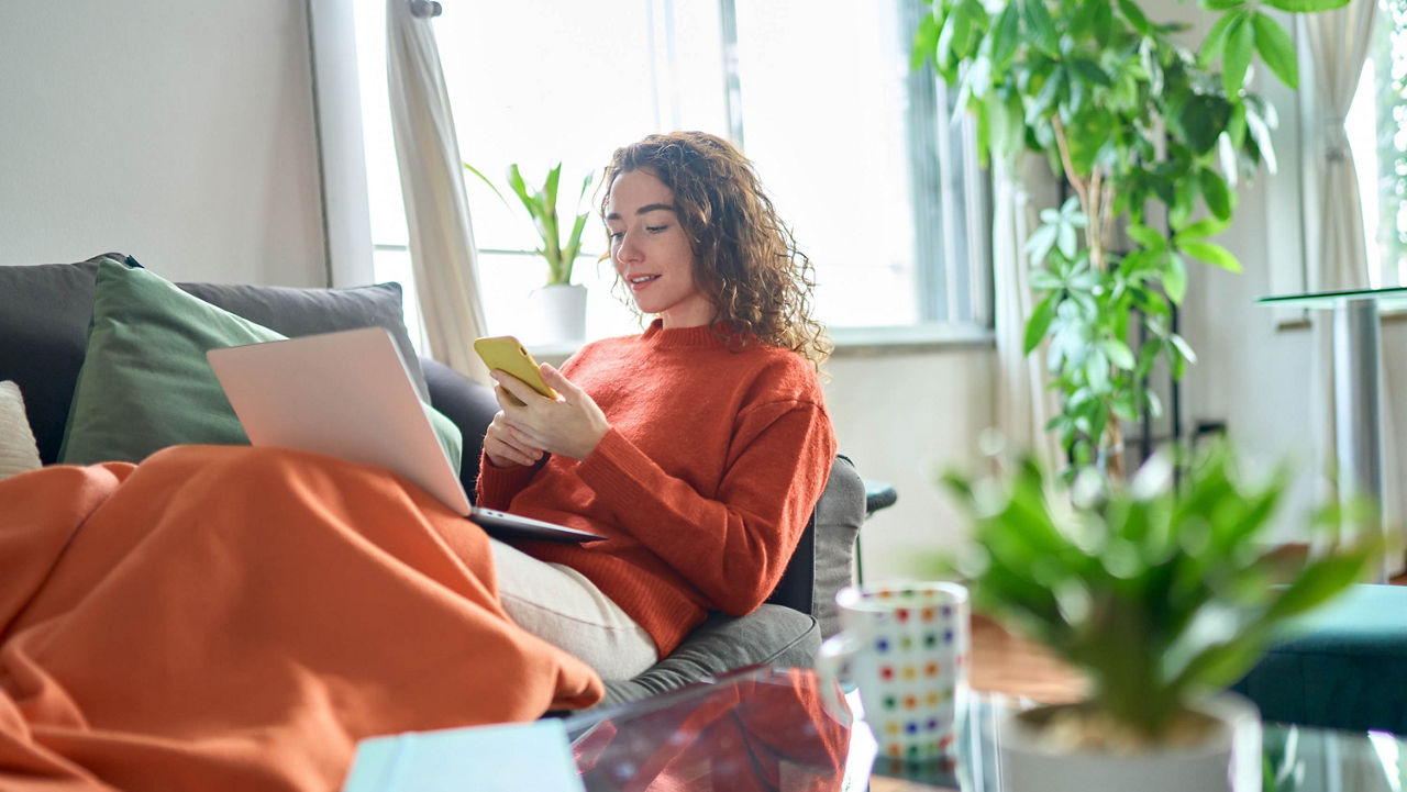 Junge Frau sitzt auf dem Sofa mit einem Laptop auf dem Schoß und einem Handy in der Hand