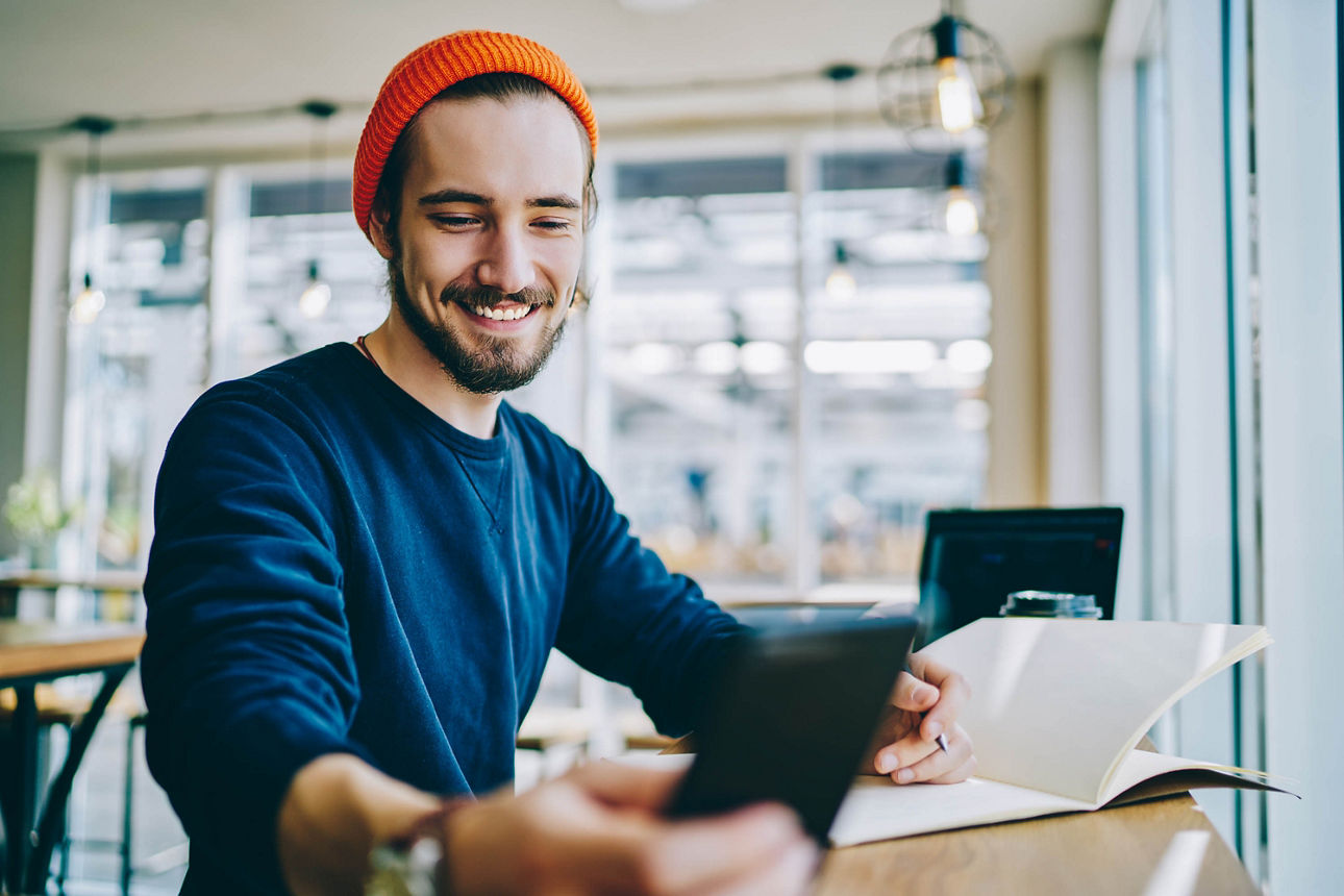 Kaukasischer männlicher Student, der mit 4g verbunden ist, um auf einem Smartphone im Internet zu surfen, während er mit einem Lehrbuch für die Ausbildung lernt, erfolgreicher Hipster, der eine Benachrichtigung auf seinem Mobiltelefon überprüft