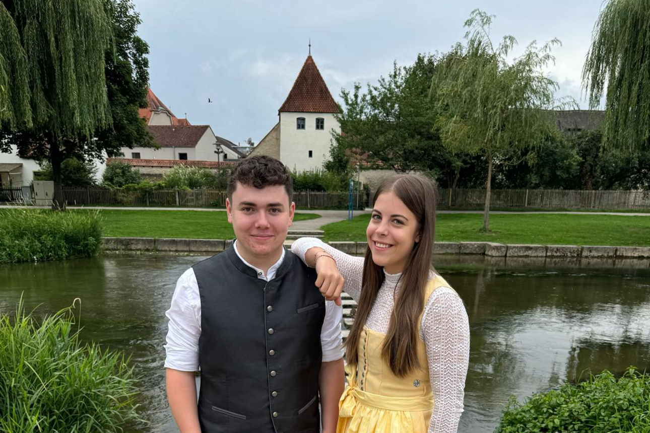 Ein junger Mann in Lederhose und Weste sowie eine junge Frau in gelbem Dirndl stehen lächelnd auf einem Weg vor einem Fluss.