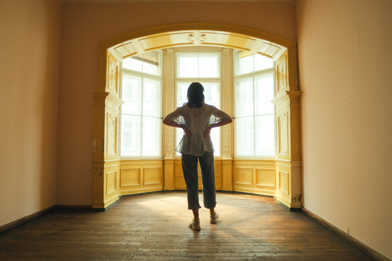 Eine Frau steht in lässigen Klamotten in einem leeren Raum vor einer Fensterfront und blickt nach draußen. Eine Frau steht in lässigen Klamotten in einem leeren Raum vor einer Fensterfront und blickt nach draußen.
