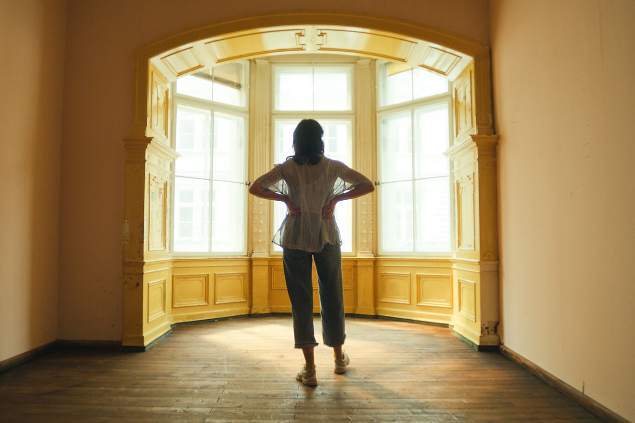 Eine Frau steht in lässigen Klamotten in einem leeren Raum vor einer Fensterfront und blickt nach draußen. Eine Frau steht in lässigen Klamotten in einem leeren Raum vor einer Fensterfront und blickt nach draußen.
