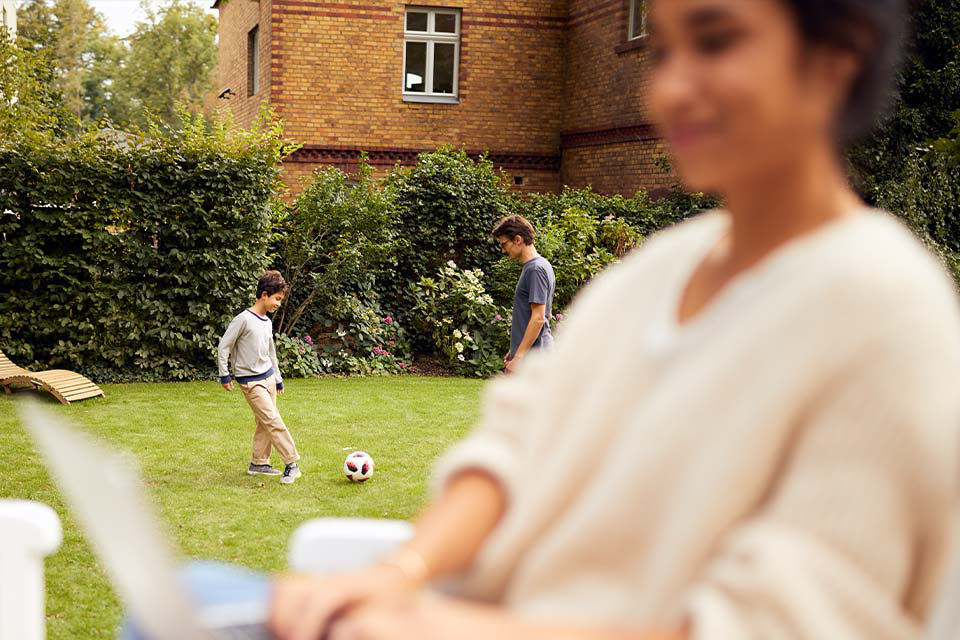 Junge Frau sitzt mit Laptop im Garten, während ein Mann und ein Kind im Hintergrund Fussball spielen