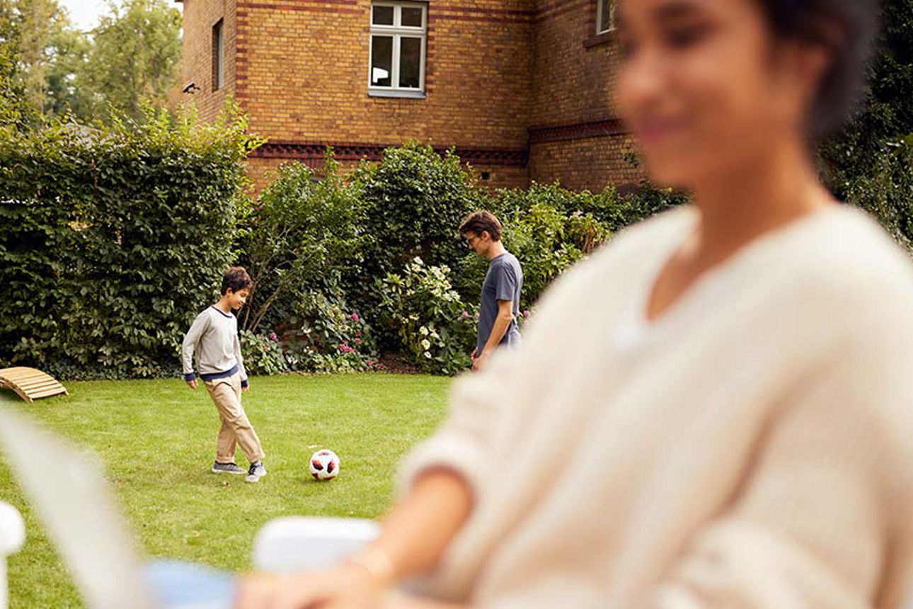 Junge Frau sitzt am Laptop im Garten und im Hintergrund spielen ein Junge und ein Mann Fußball