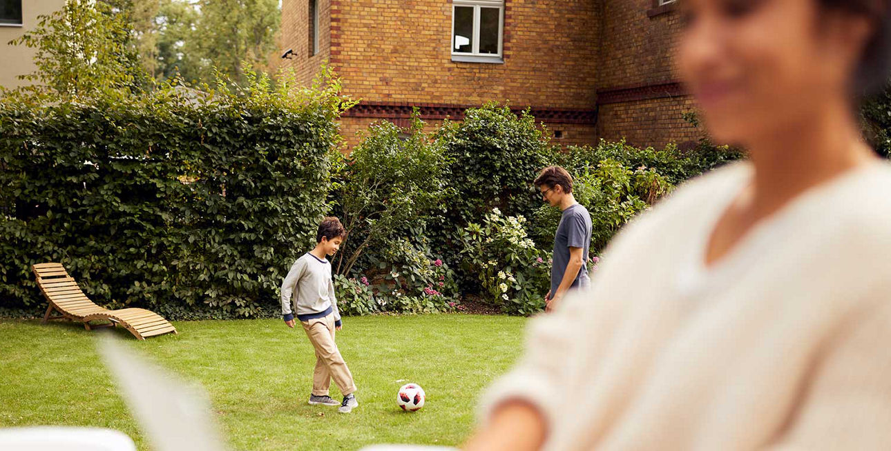 Junge Frau sitzt mit Laptop im Garten, während ein Mann und ein Kind im Hintergrund Fußball spielen