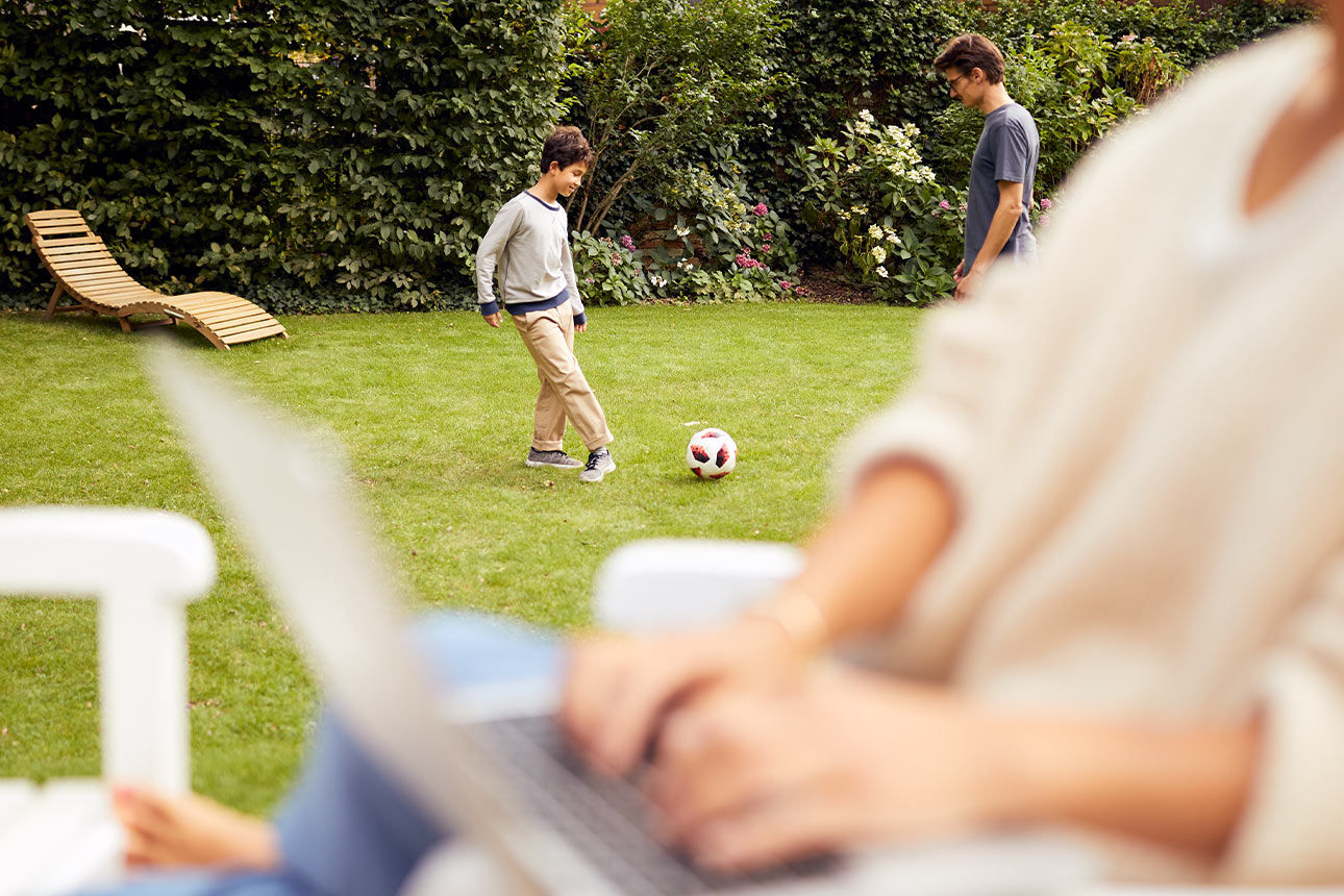 Junge Frau sitzt mit Laptop im Garten, während ein Mann und ein Kind im Hintergrund Fußball spielen