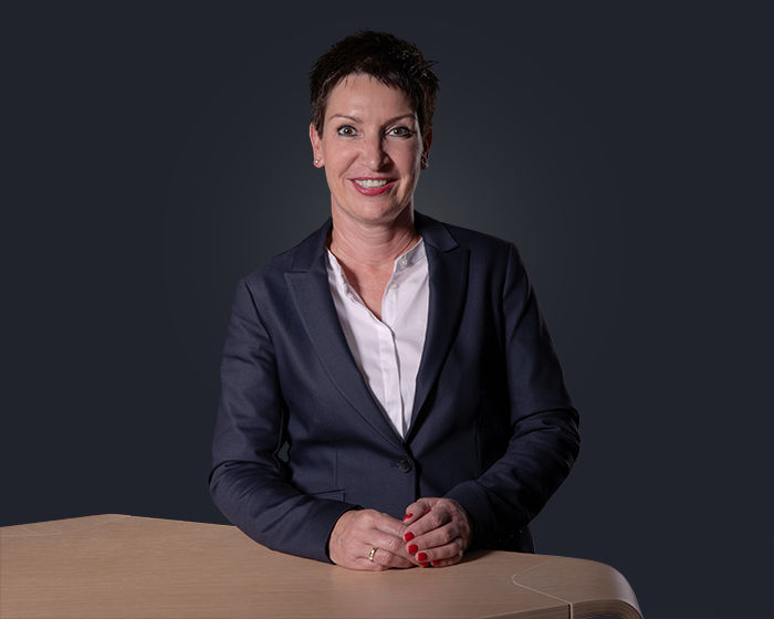 Porträt von Heike Rüther-Tietze, lächelnd, an einem Schreibtisch sitzend, dunkelblauer Hintergrund