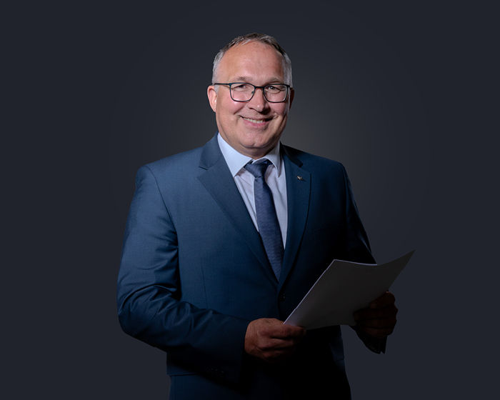 Porträt von Michael Brenke lächelnd und mit Papieren in der Hand vor dunkelblauem Hintergrund
