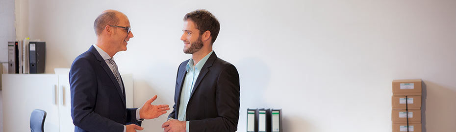 Zwei Männer in Anzügen stehen in einem Büro und führen ein Gespräch. 