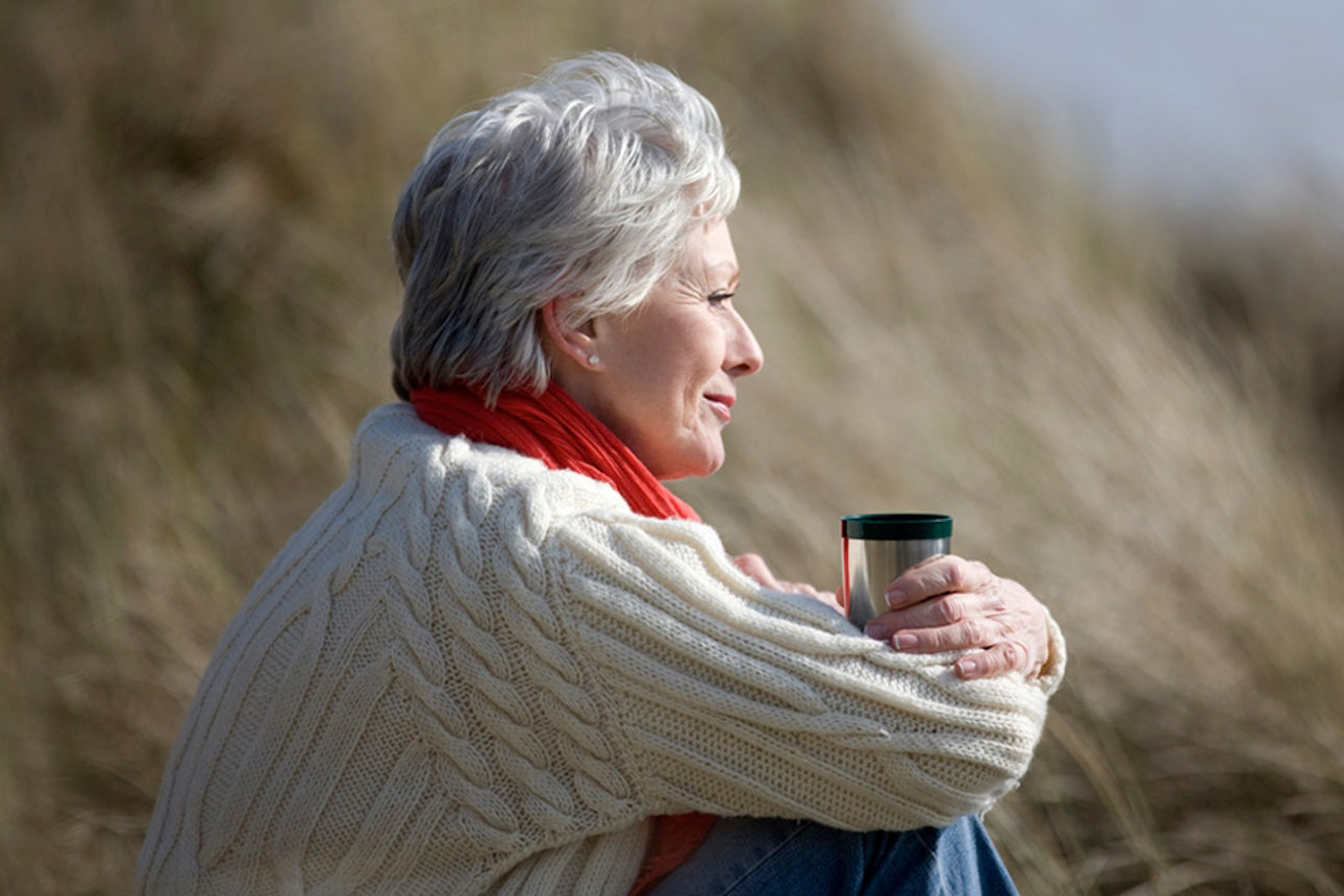 Ältere Frau sitzt gedankenverloren mit verschränkten Armen und einer Tasse in der Hand