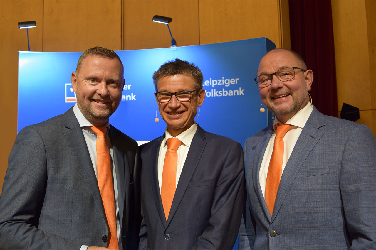 3 Herren stehen vor Werbewand und lächeln