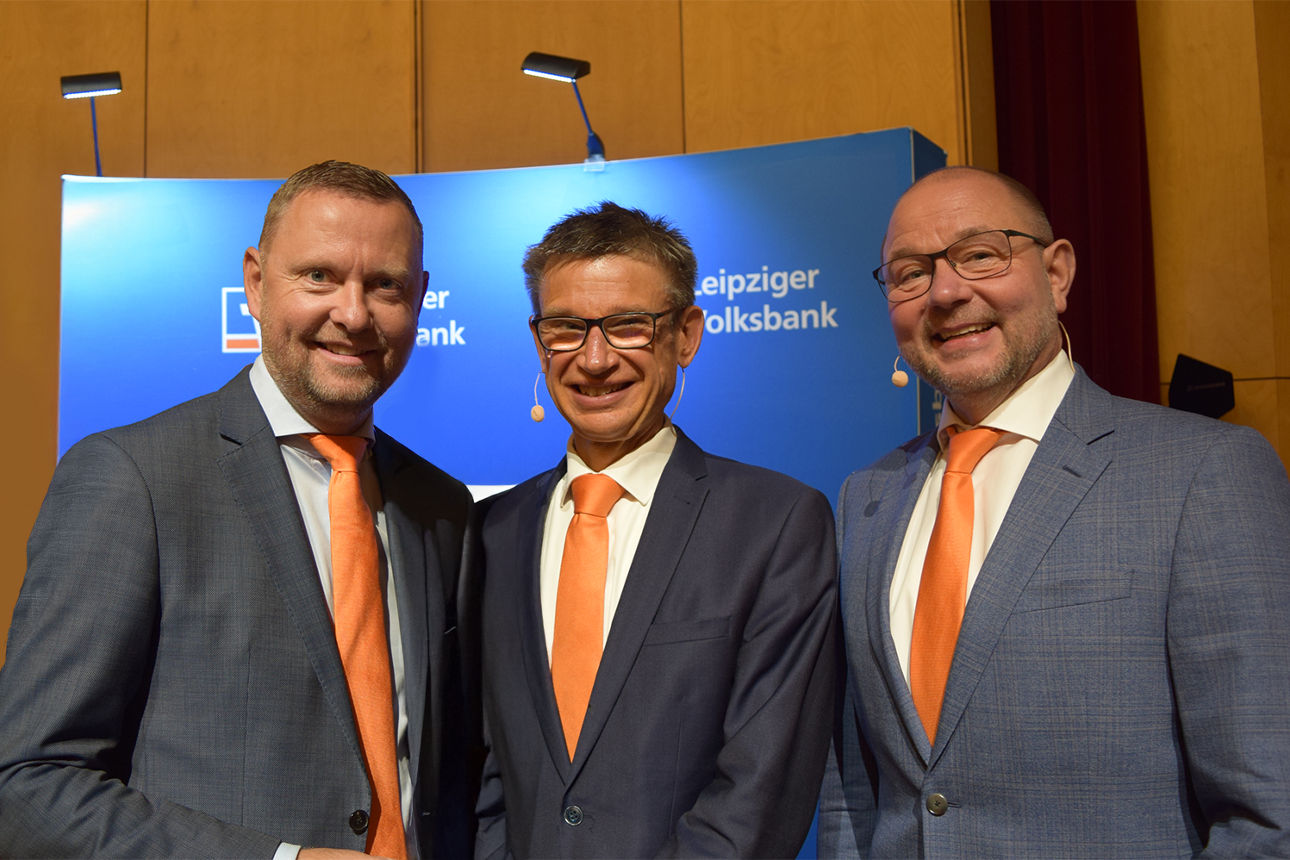 3 Herren stehen vor Werbewand und lächeln