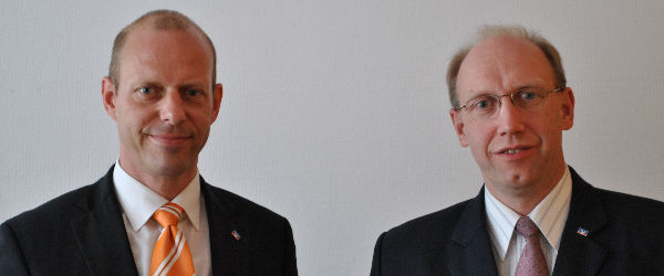 Zwei Männer in Anzügen, einer mit orangefarbener Krawatte, der andere mit kastanienbrauner Krawatte, schauen in die Kamera