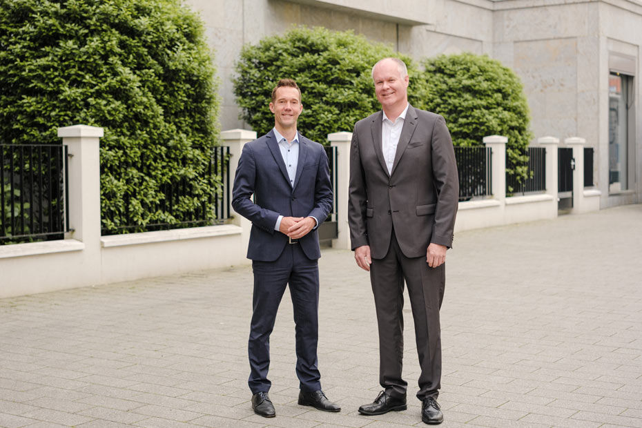 Zwei Männer in Anzügen. Sie stehen im Freien, wahrscheinlich neben einem Gebäude