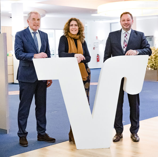 Drei Personen stehen in einer professionellen Umgebung und posieren vor einem großen weißen Buchstaben „V“.
