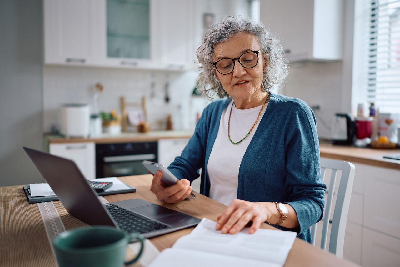 Frau am Laptop, die auf ihr Smartphone schaut