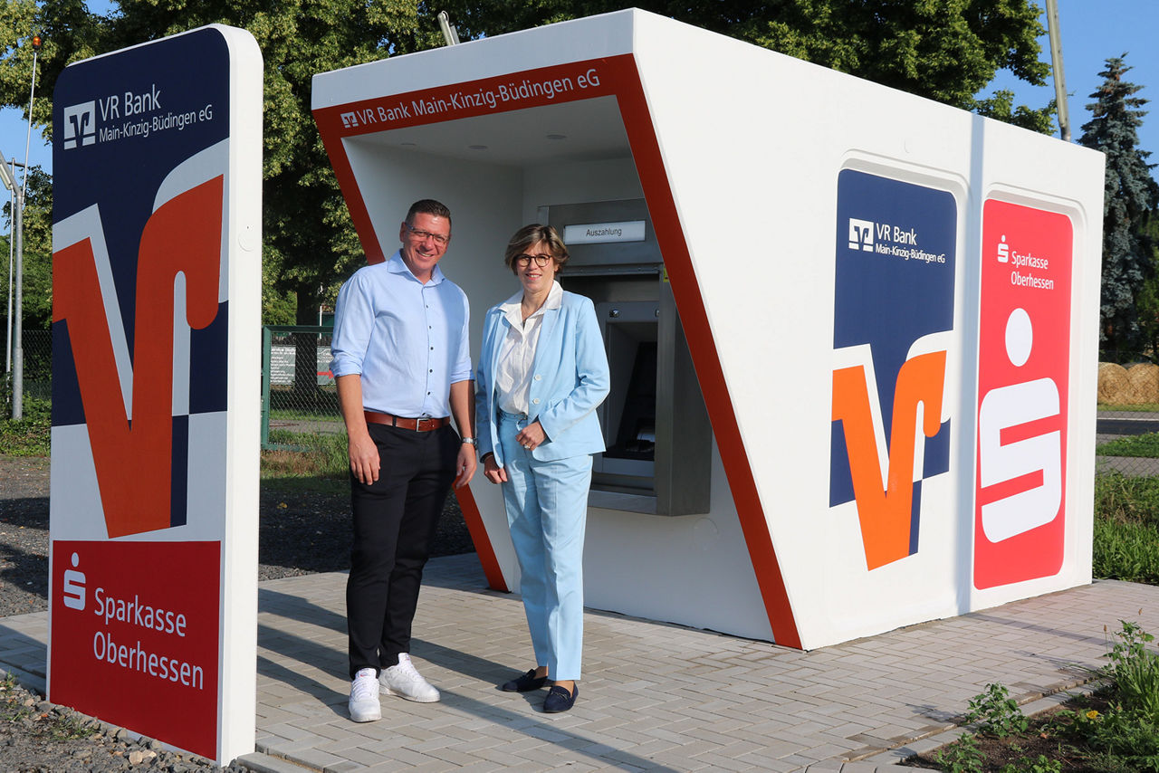 Sabine Haas und Christoph Appel im Business-Outfit vor hellem Hintergrund, lächelnd am weißen Tresen mit VR-Logo.