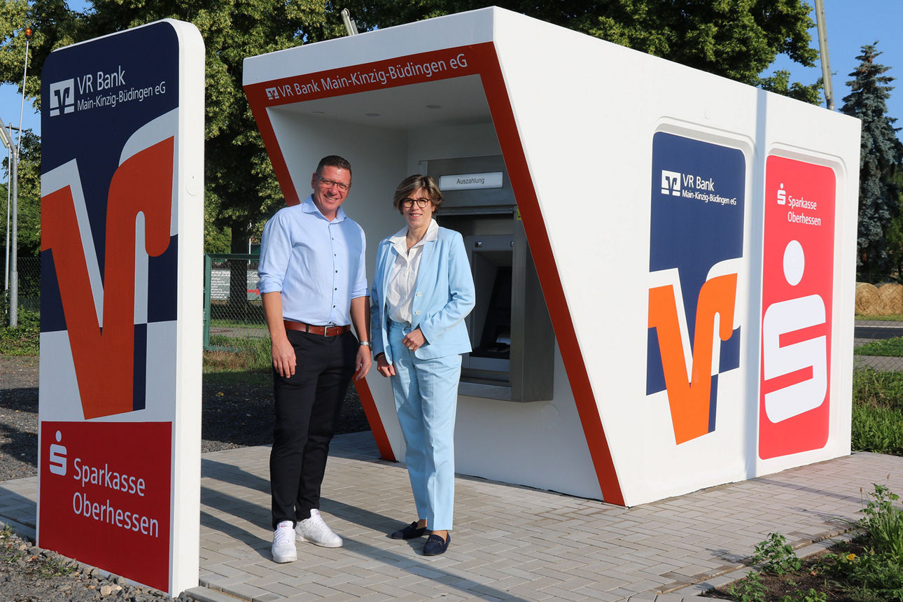 Sabine Haas und Christoph Appel im Business-Outfit vor hellem Hintergrund, lächelnd am weißen Tresen mit VR-Logo.