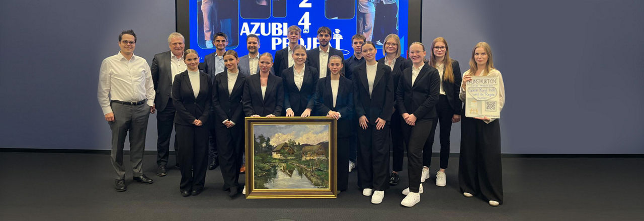 Der Azubi-Jahrgang 2024 steht gemeinsam mit vier Verantwortlichen vor einer blauen Wand und hält ein Bild der Auktion in der Hand. 