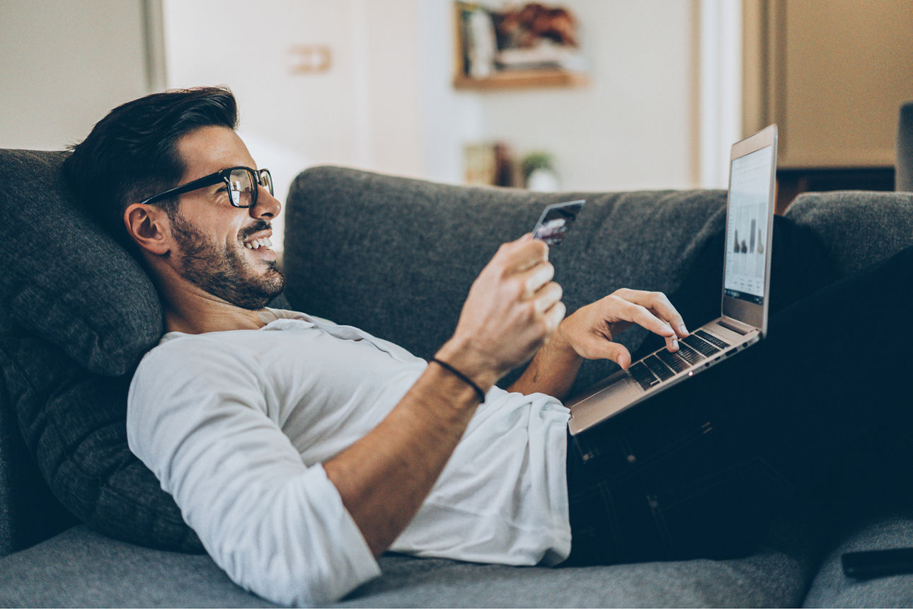 Ein Mann liegt auf der Couch mit seinem Laptop und hält eine Kreditkarte in der Hand