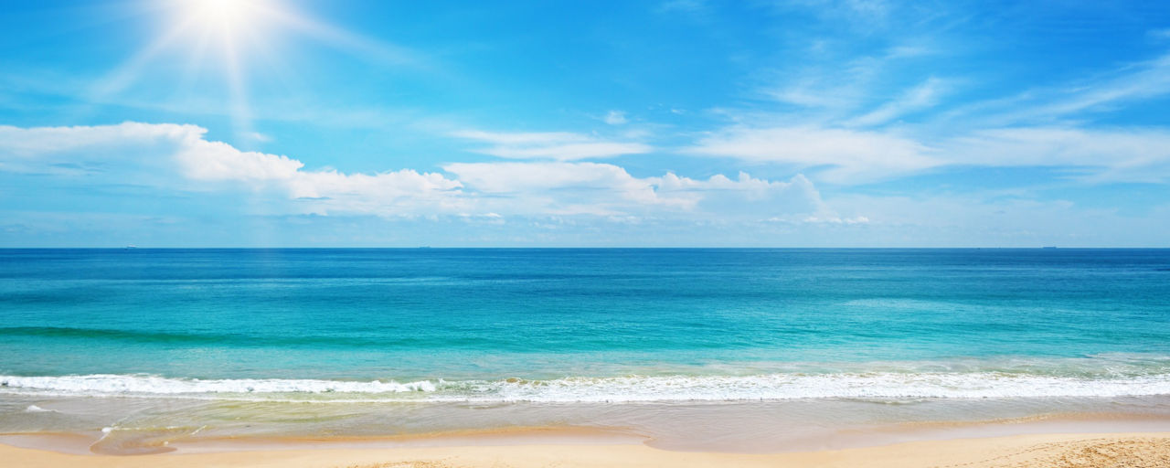 Blick über einen Sandstrand auf türkisblaues Meer und Sonne