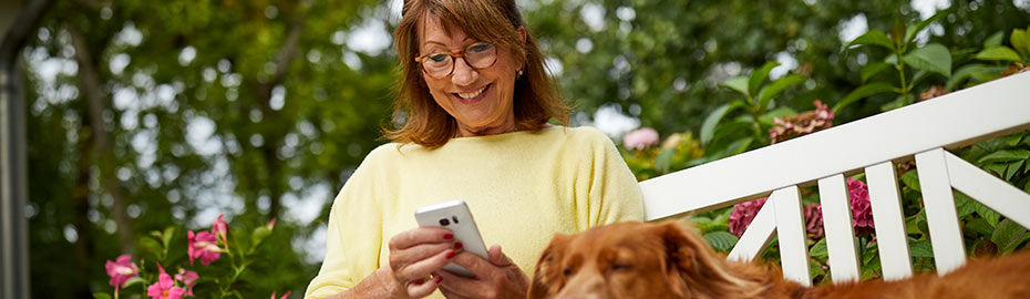 Frau mittleren Alters mit Brille, die lächelnd auf ihr Telefon schaut, während ein Hund neben ihr sitzt