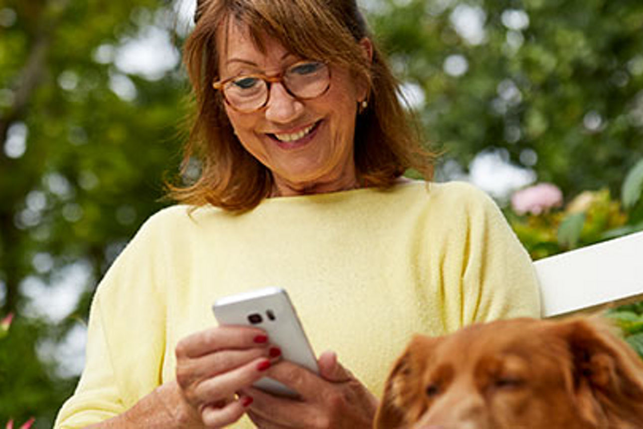Frau mittleren Alters mit Brille, die lächelnd auf ihr Telefon schaut, während ein Hund neben ihr sitzt