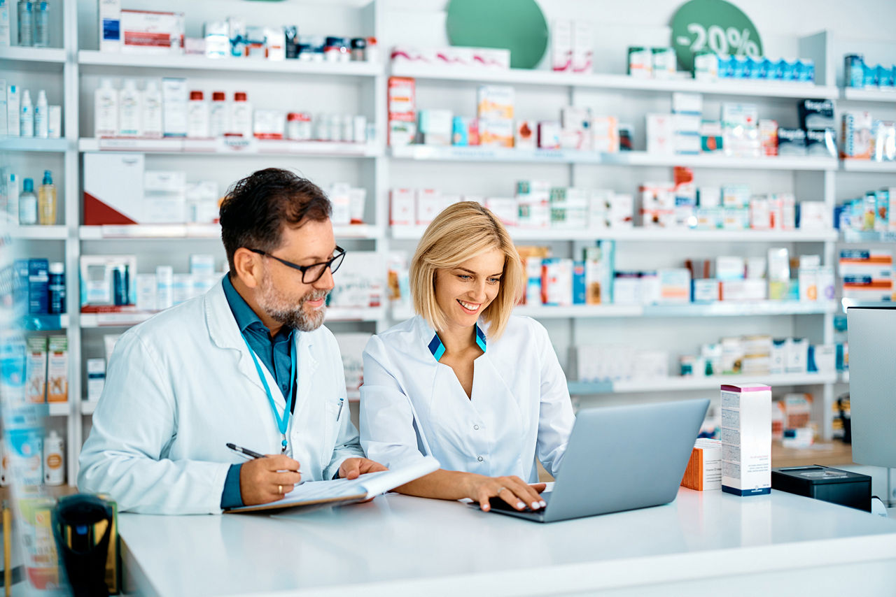 Apothekerin sitzt in der Apotheke am Laptop, ihr Kollege macht sich Notizen. 
