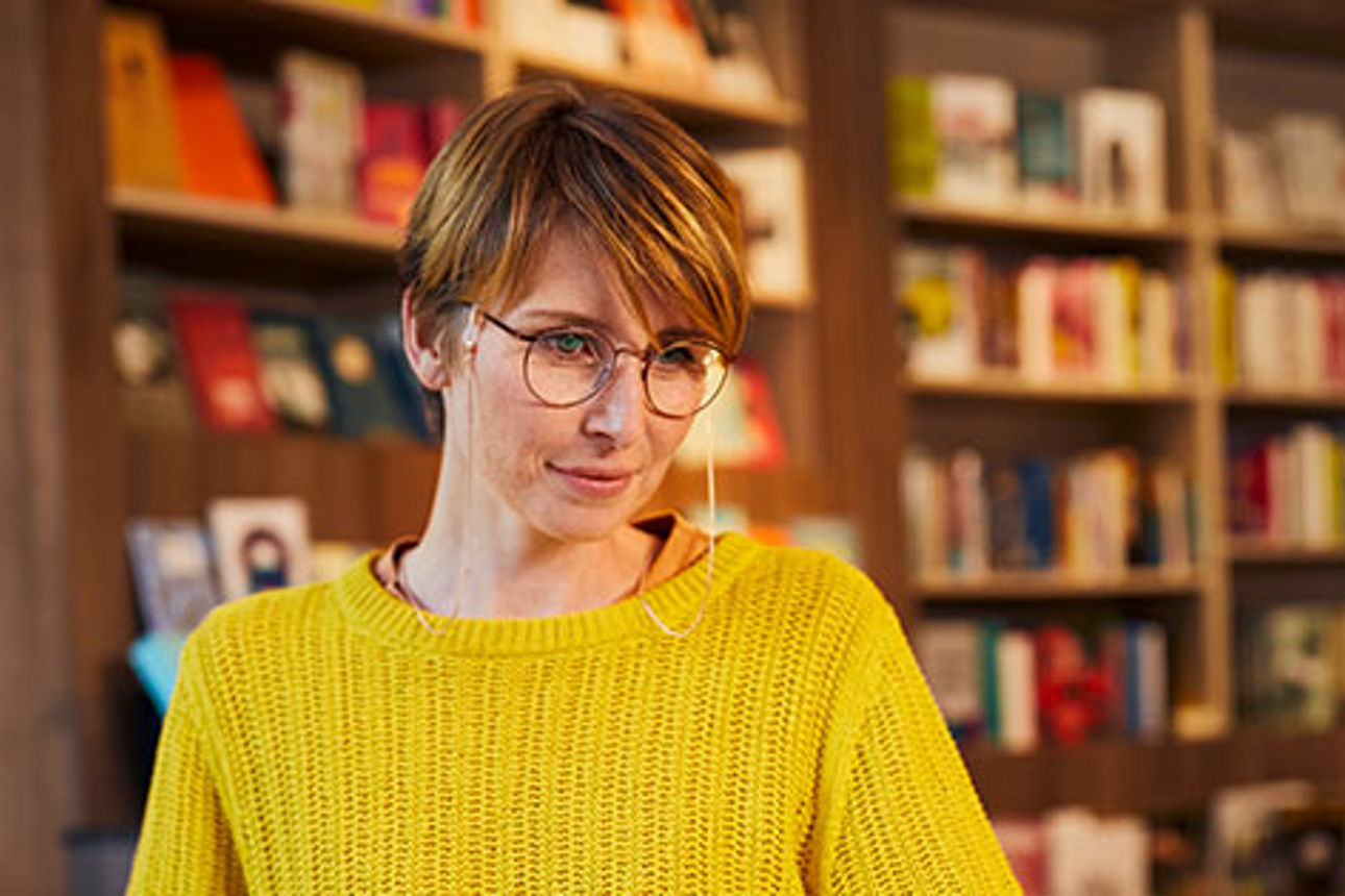 Eine Frau in einem gelben Pullover steht selbstbewusst vor einem Bücherregal