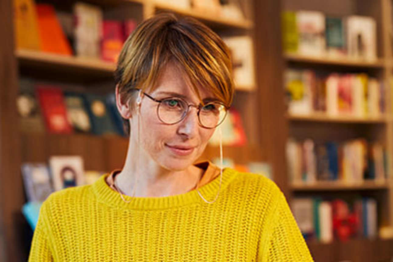 Eine Frau mit Brille und gelbem Pullover vor einer Bibliothek mit Büchern