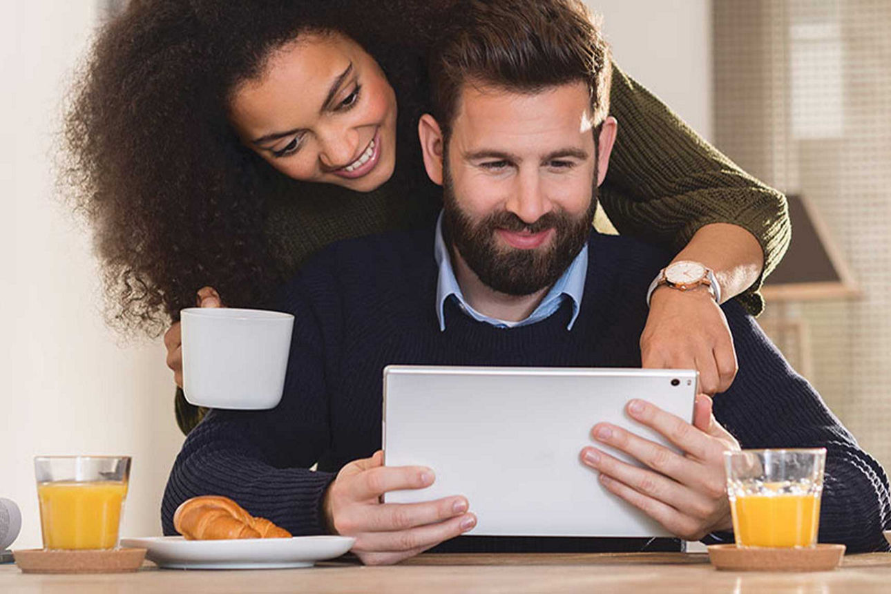 Ein Mann sitzt am Tisch und hat ein Ipad in der Hand, hinter ihm steht eine Frau mit Kaffeetasse und schaut auf das Ipad