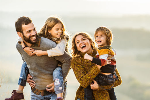 Familie mit zwei Kindern macht einen Spaziergang. Die Kinder werden von den Eltern auf Arm und Rücken getragen.