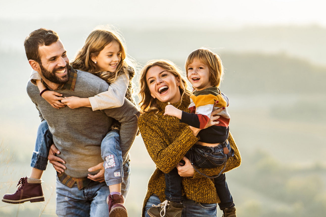 Familie mit zwei Kindern macht einen Spaziergang. Die Kinder werden von den Eltern auf Arm und Rücken getragen.