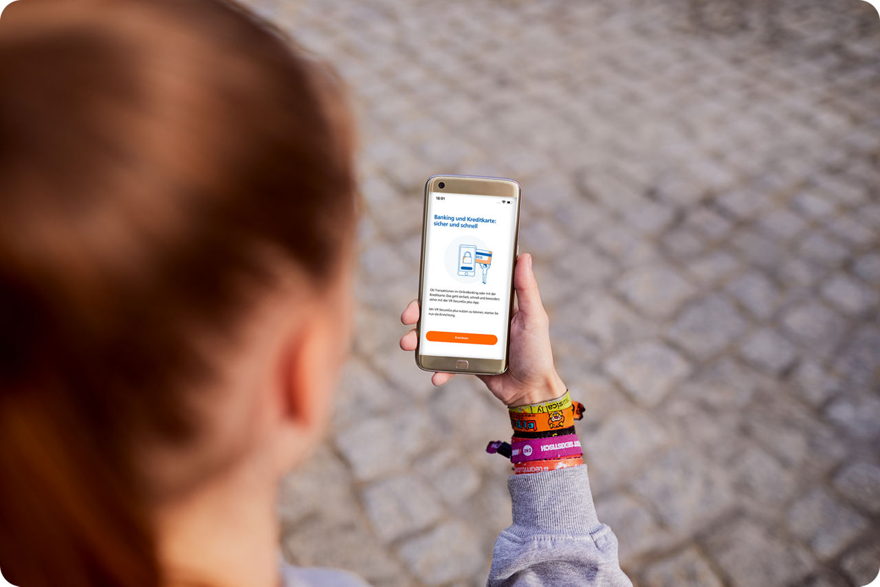 Eine junge Frau hält ein Smartphone in der Hand, auf dem der Einrichtungs-Startbildschirm der VR SecureGo plus App zu sehen ist.