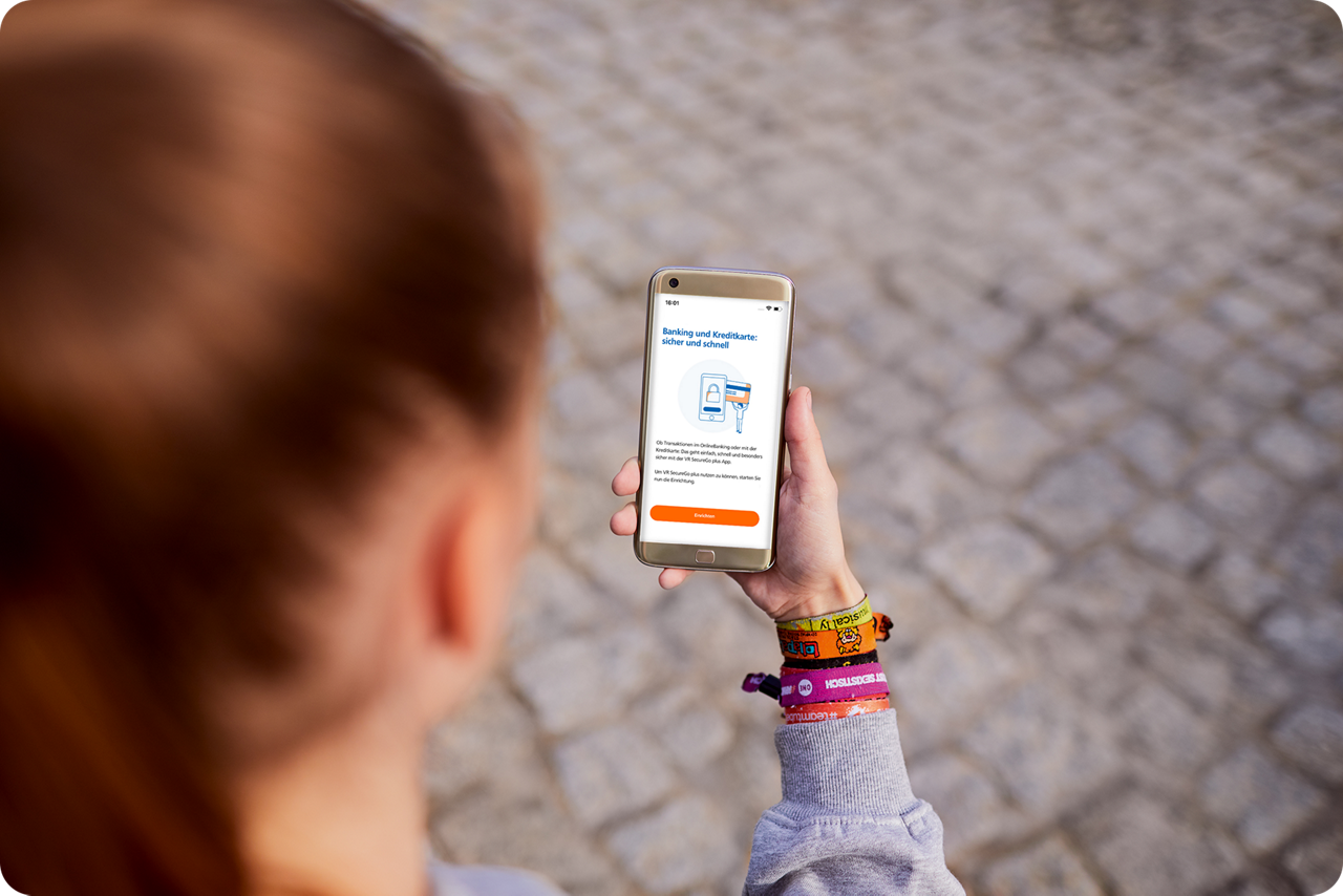 Eine junge Frau hält ein Smartphone in der Hand, auf dem der Einrichtungs-Startbildschirm der VR SecureGo plus App zu sehen ist.