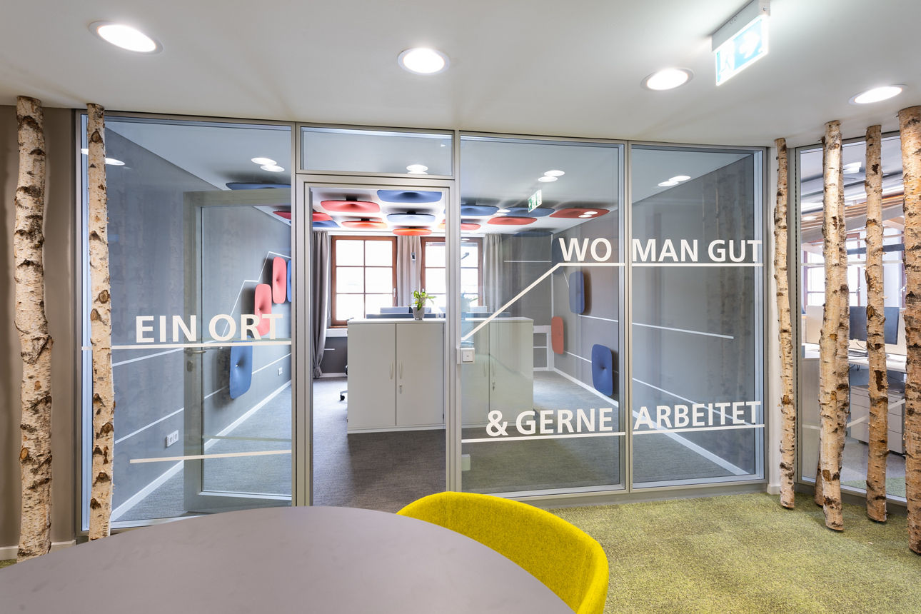 Gläserne Bürotüre mit Aufschrift "Ein Ort wo man gut und gerne arbeitet" mit Blick in modernes Büro, seitlich Birken Birkenst