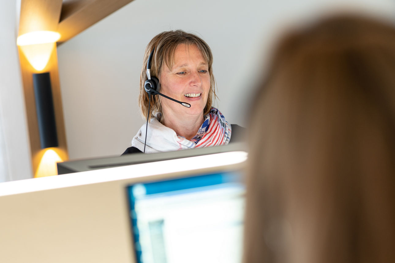 Frau hinter PC-Bildschirm im Büro mit Kopfhörern; im Vordergrund verschwommen ein Teilasuschnitt von Bildschirm und Kopf